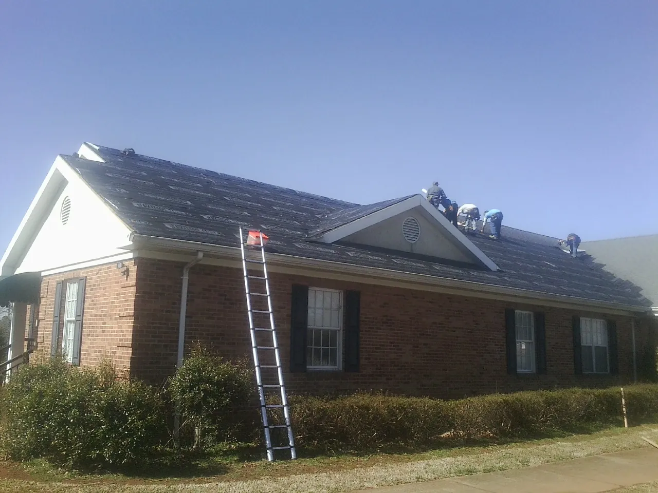 Crew tearing shingles off a brick ranch roof with a ladder set — Metal Roof Repair work by our crew in Brodhead