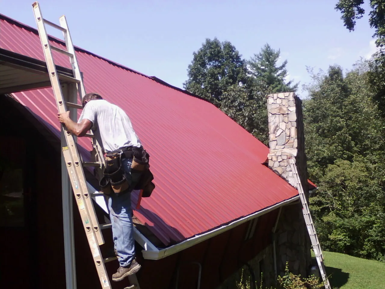 Roofer on a ladder beside a red metal roof and stone chimney — Soffit & Fascia Repair by Roofing Brothers in Brodhead